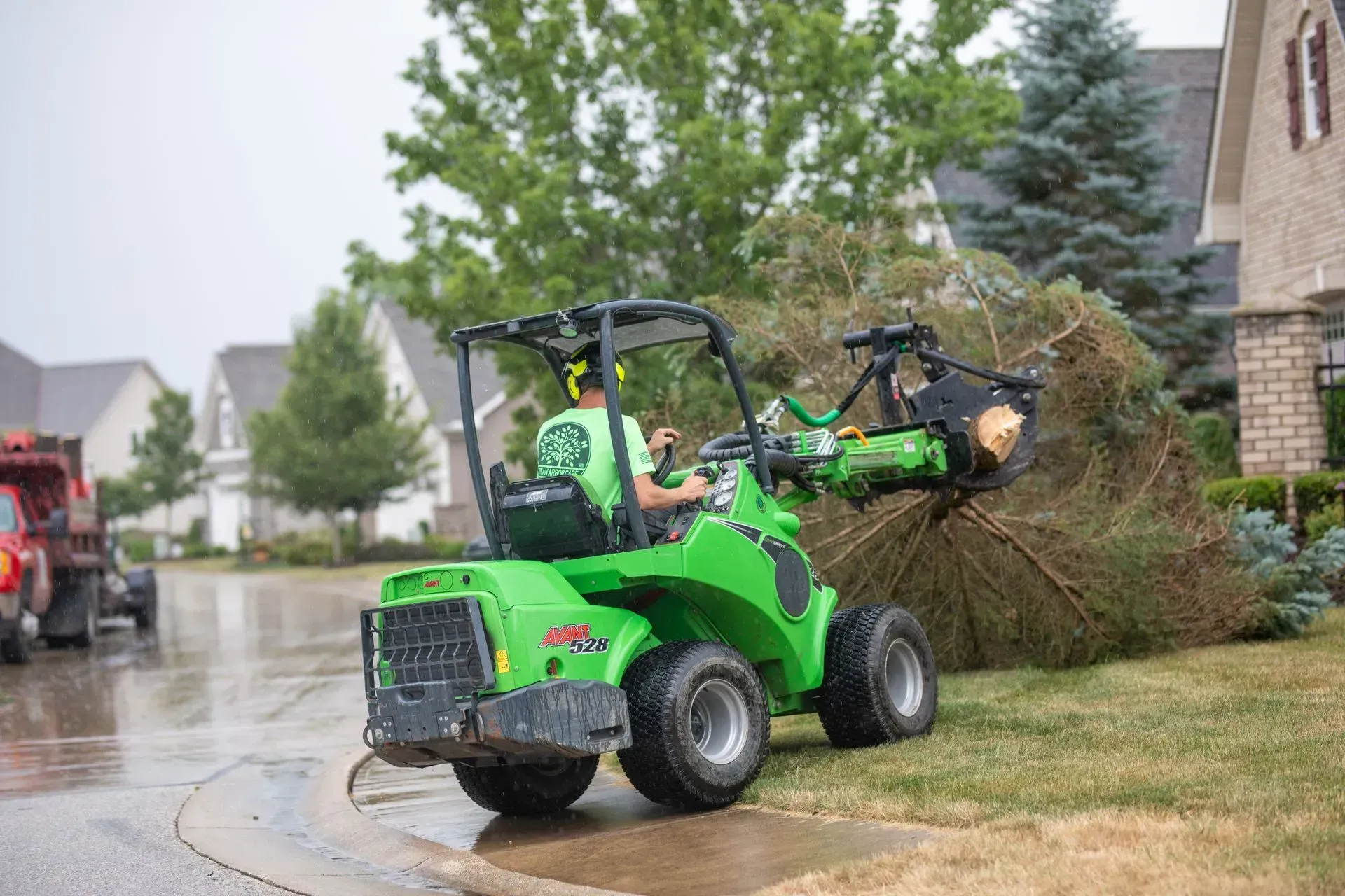 A green compact loader with a tree trunk in its arms, being operated by a person on a suburban street.