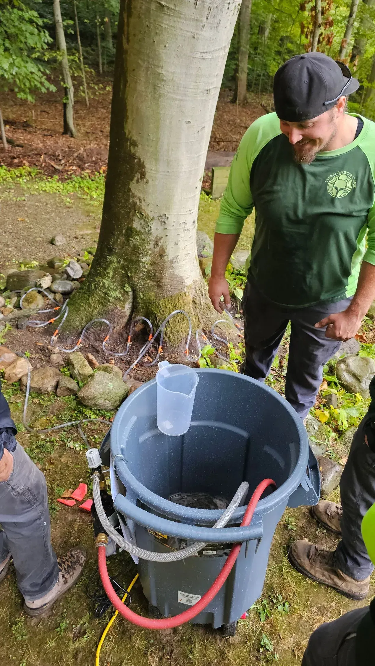 Man smiles near a large bucket with hoses, possibly using equipment in a wooded setting.
