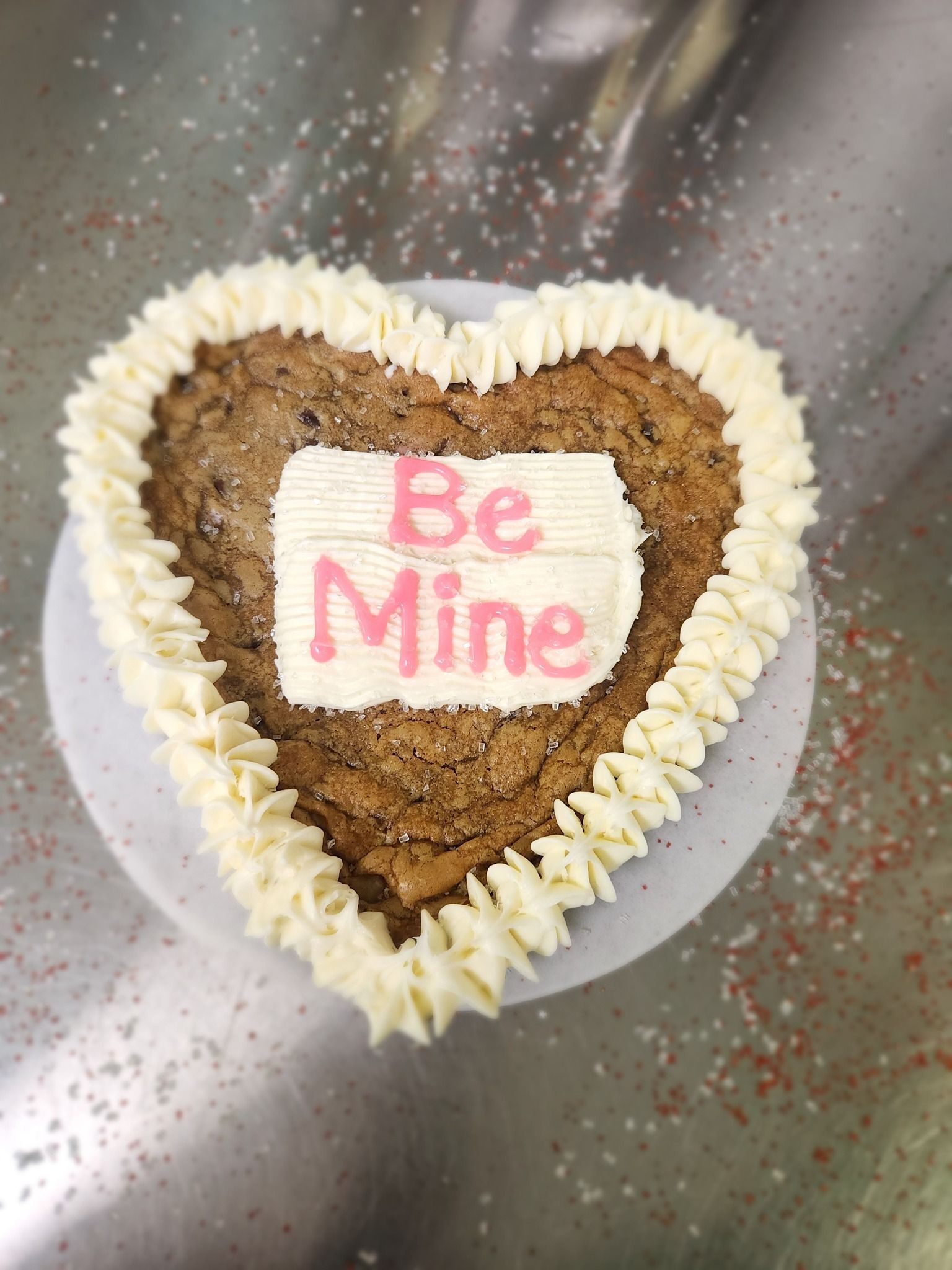 Heart-shaped cake with white frosting and pink “Be Mine” message on a silver surface