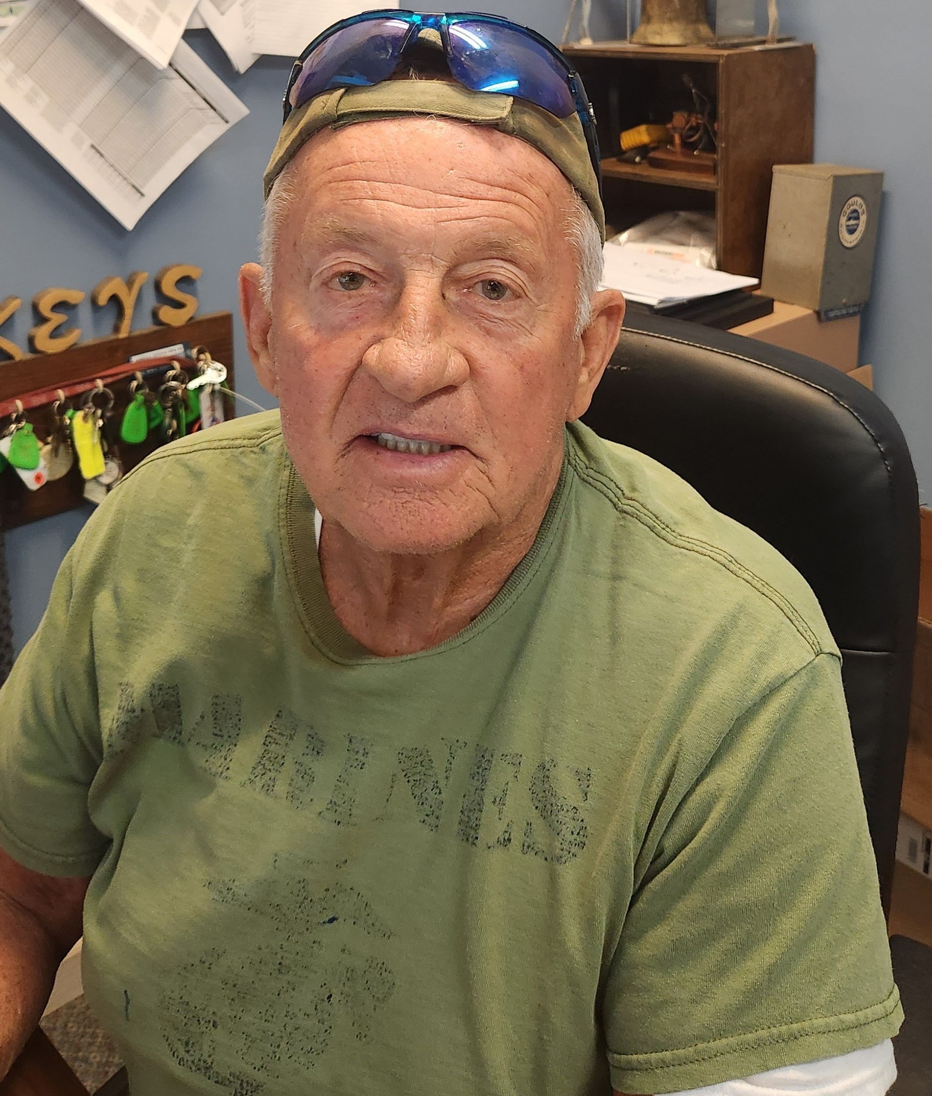 A Plumber with light skin, wearing a green shirt and blue sunglasses on his head, sits at a desk getting the work order for the day.