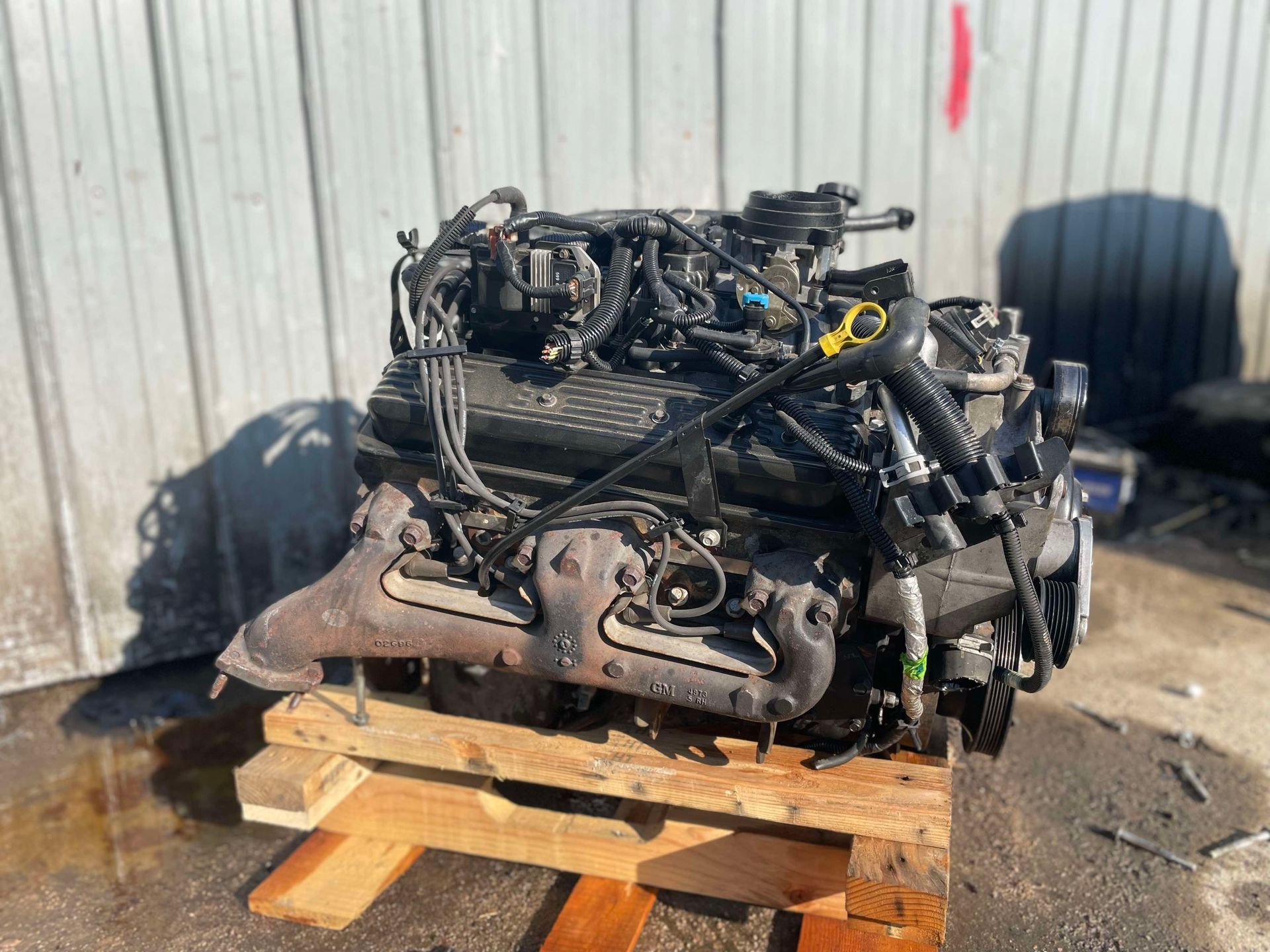 Engine on a wooden pallet; black and silver with various wires and hoses. Outdoors.