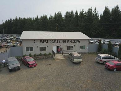 All West Coast Auto Wrecking building with parked cars.  Junkyard in the background, trees, and overcast sky.