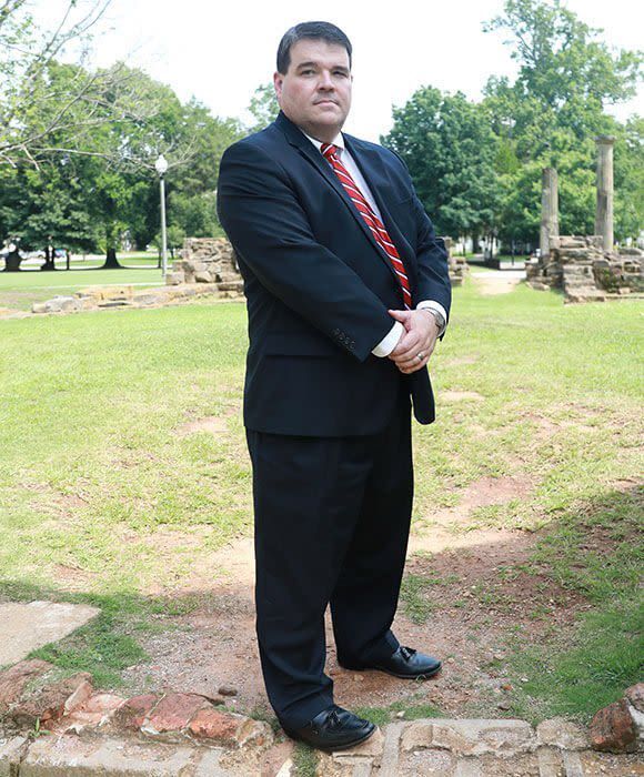 A person in a dark suit and red tie stands with hands clasped in front of them in a park with historic brick ruins.