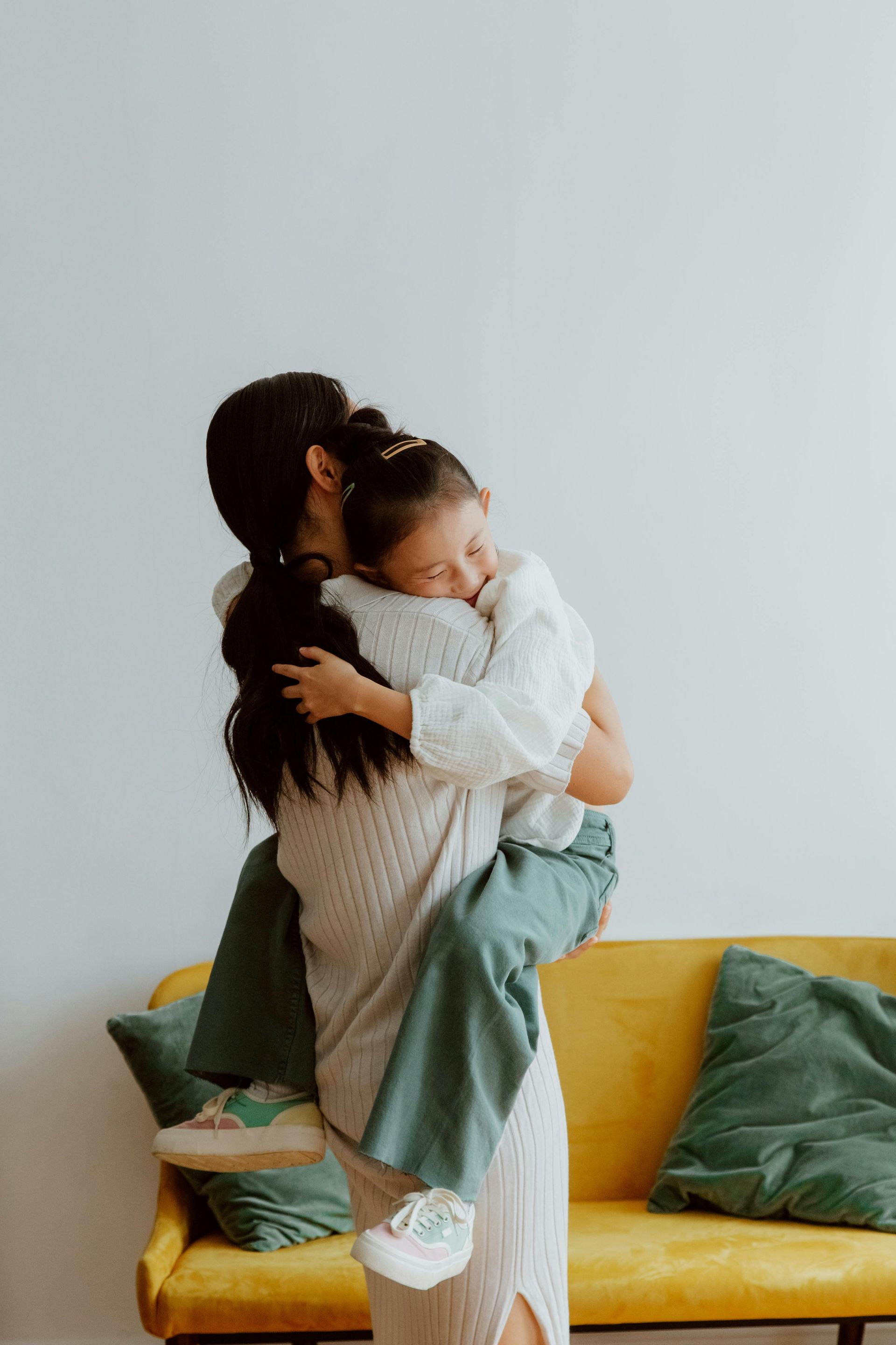 A person in a beige sweater and green pants hugs a child, both standing in front of a yellow sofa against a white wall.