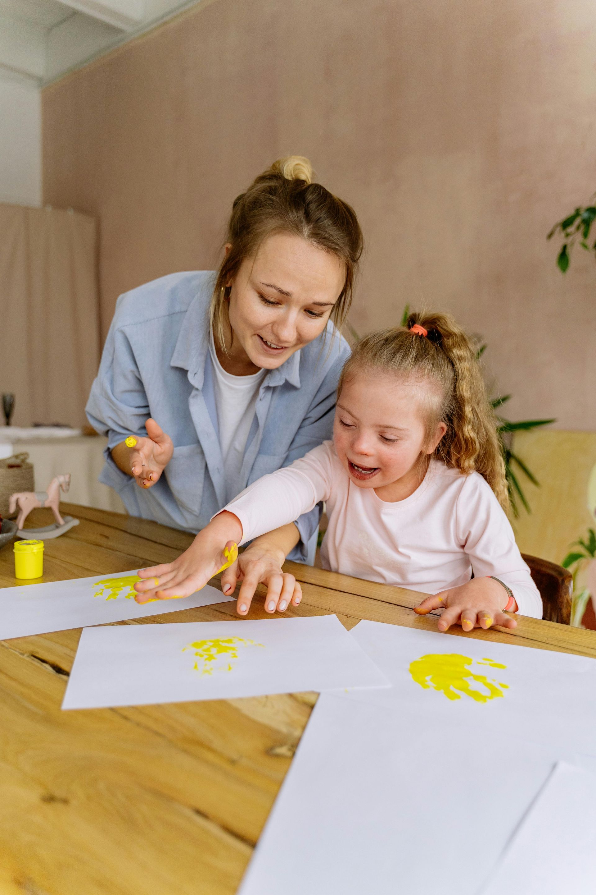 A person and a child joyfully creating handprint art with yellow paint on paper at a wooden table.