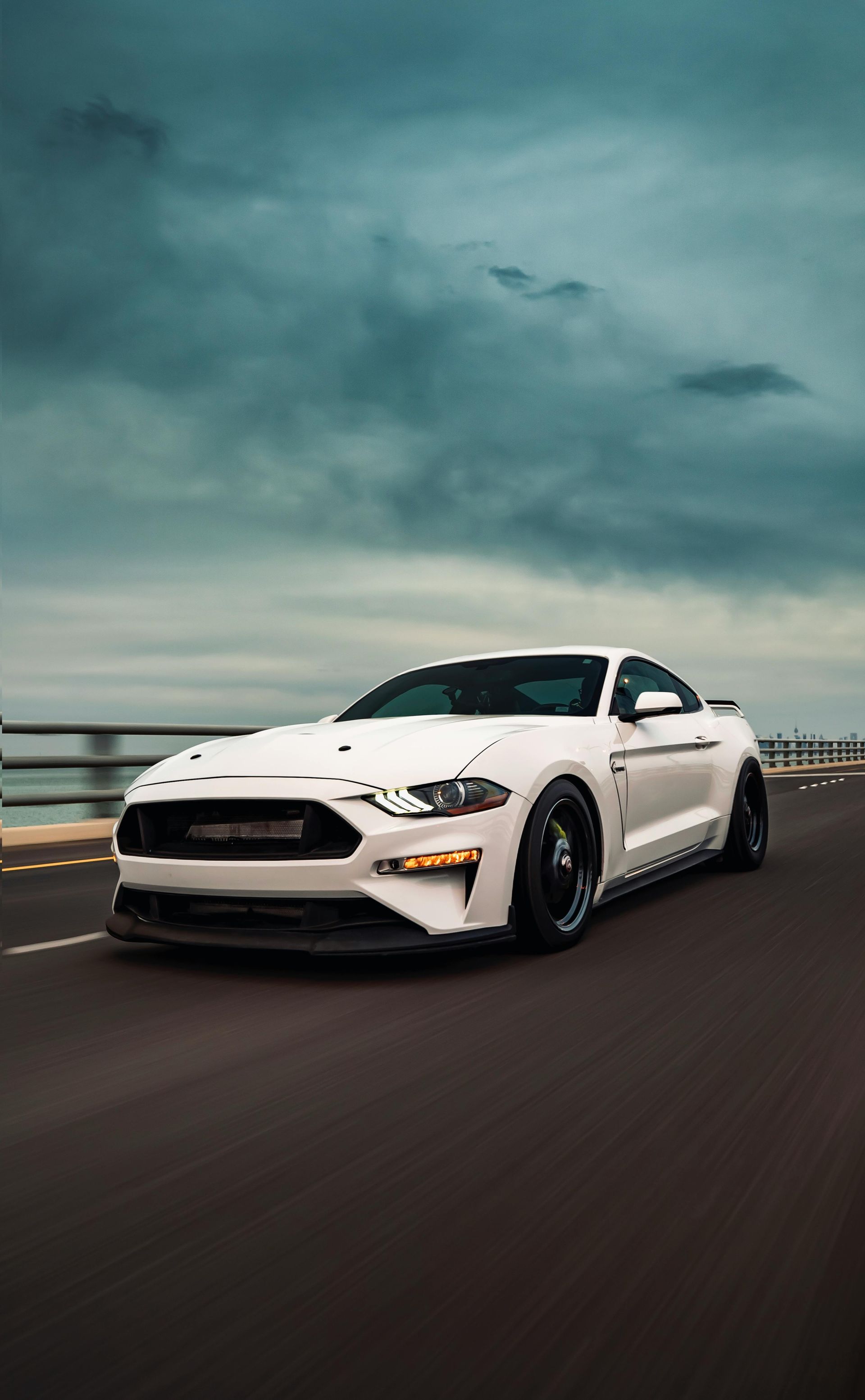 A white Ford Mustang driving on a road under a dramatic, cloudy sky.
