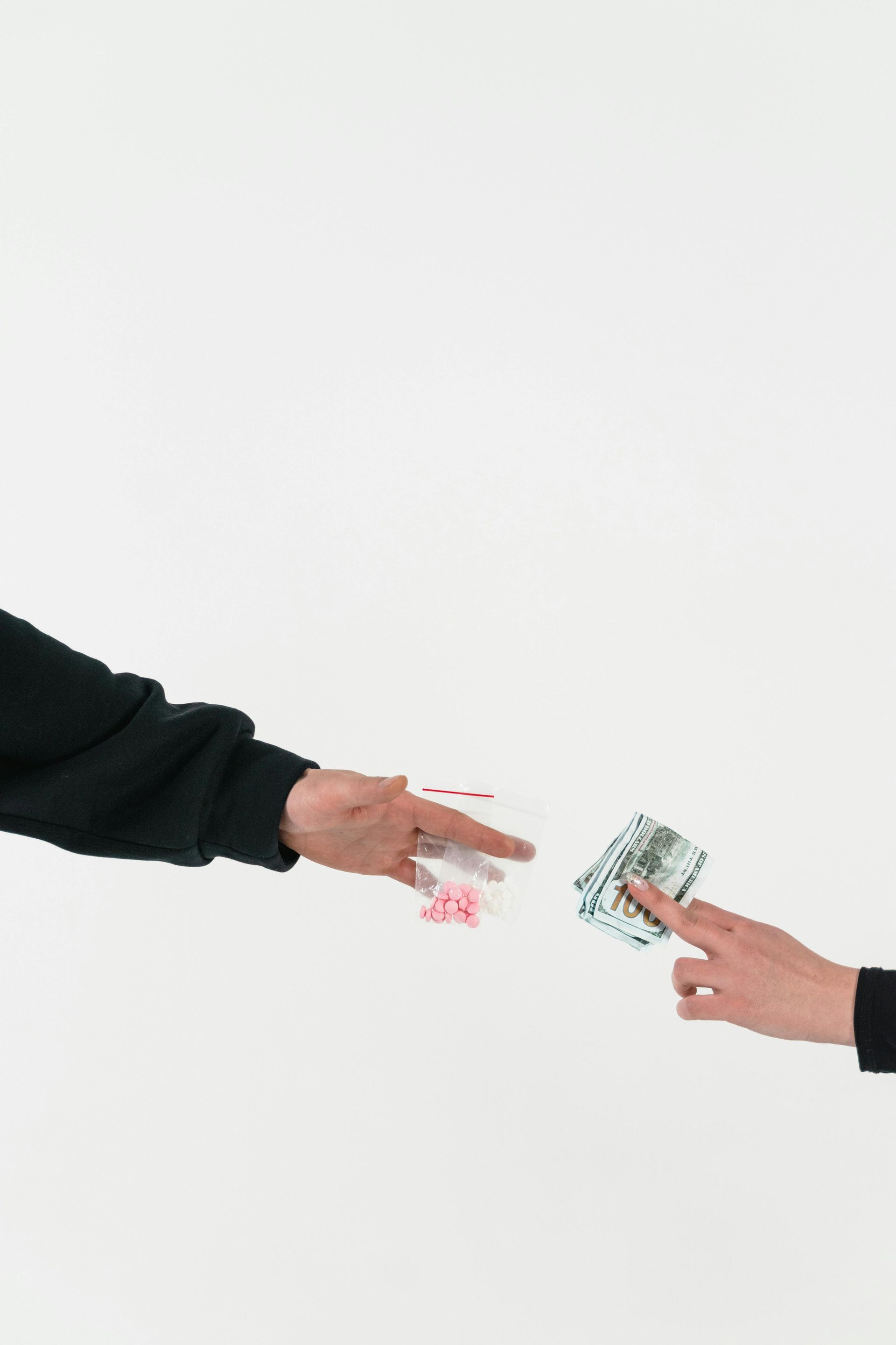 Two hands reaching toward each other, one offering a small plastic bag with pink pills and the other holding cash.