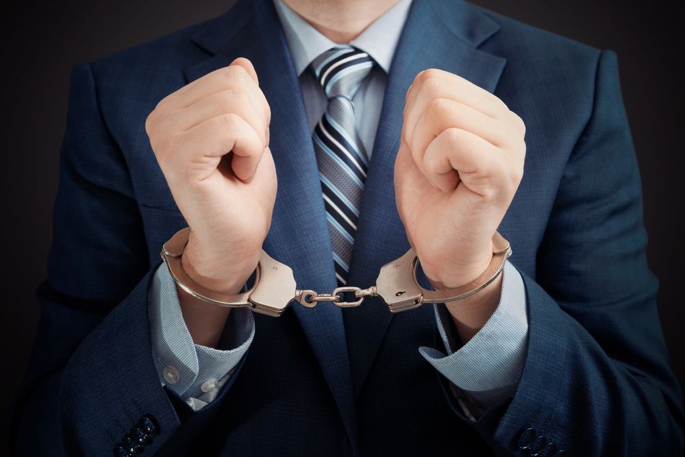A person in a business suit and striped tie with their wrists handcuffed together in front of them against a dark background.