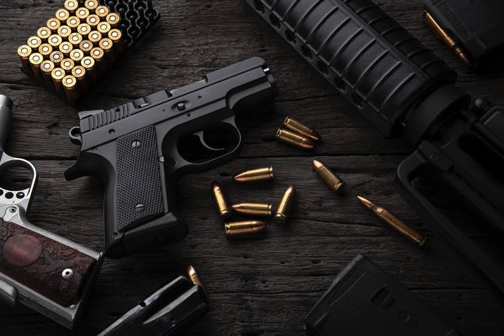 A semi-automatic pistol, rifle parts, magazines, and scattered ammunition arranged on a dark, rustic wooden surface.