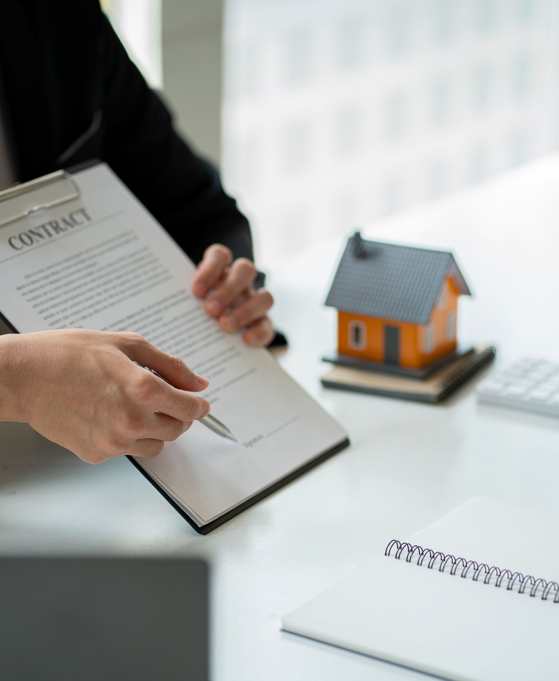 Hands reviewing a contract beside a small house model and notebook on a desk