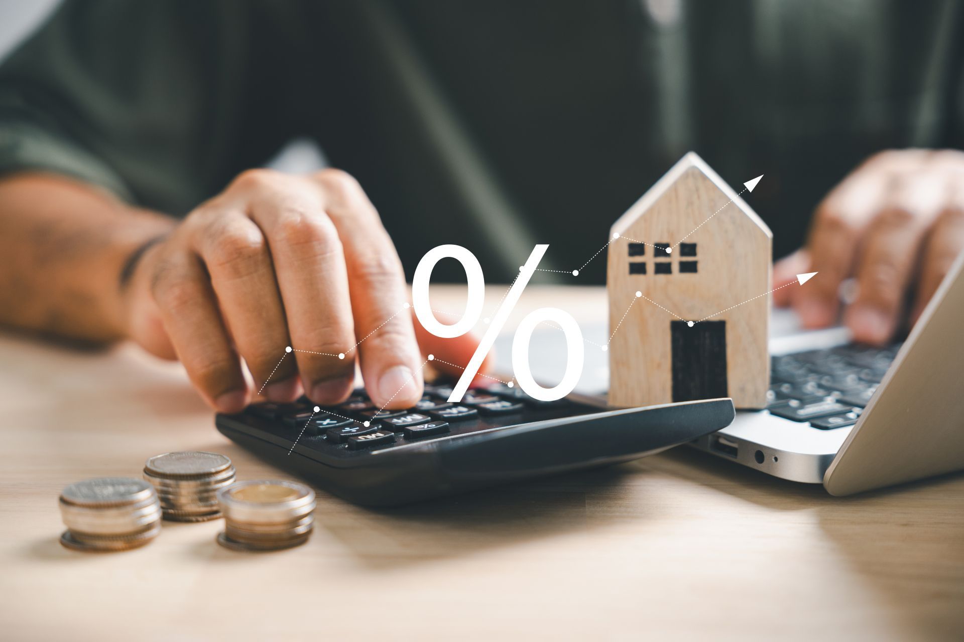 Hands typing on calculator with house model, percent symbol, and coins beside a laptop.