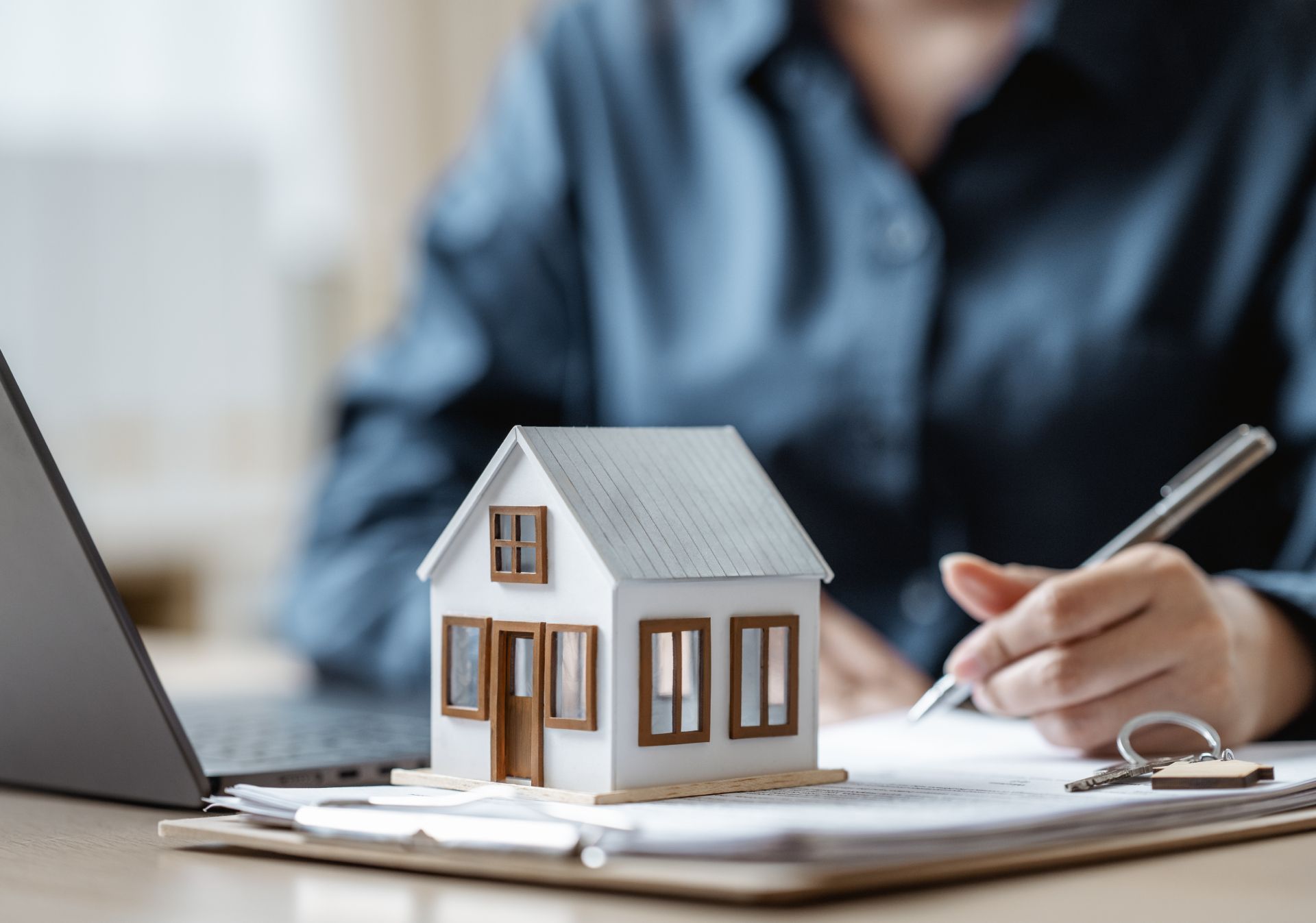 Miniature house on a desk beside a laptop and notebook, with a person writing in the background.
