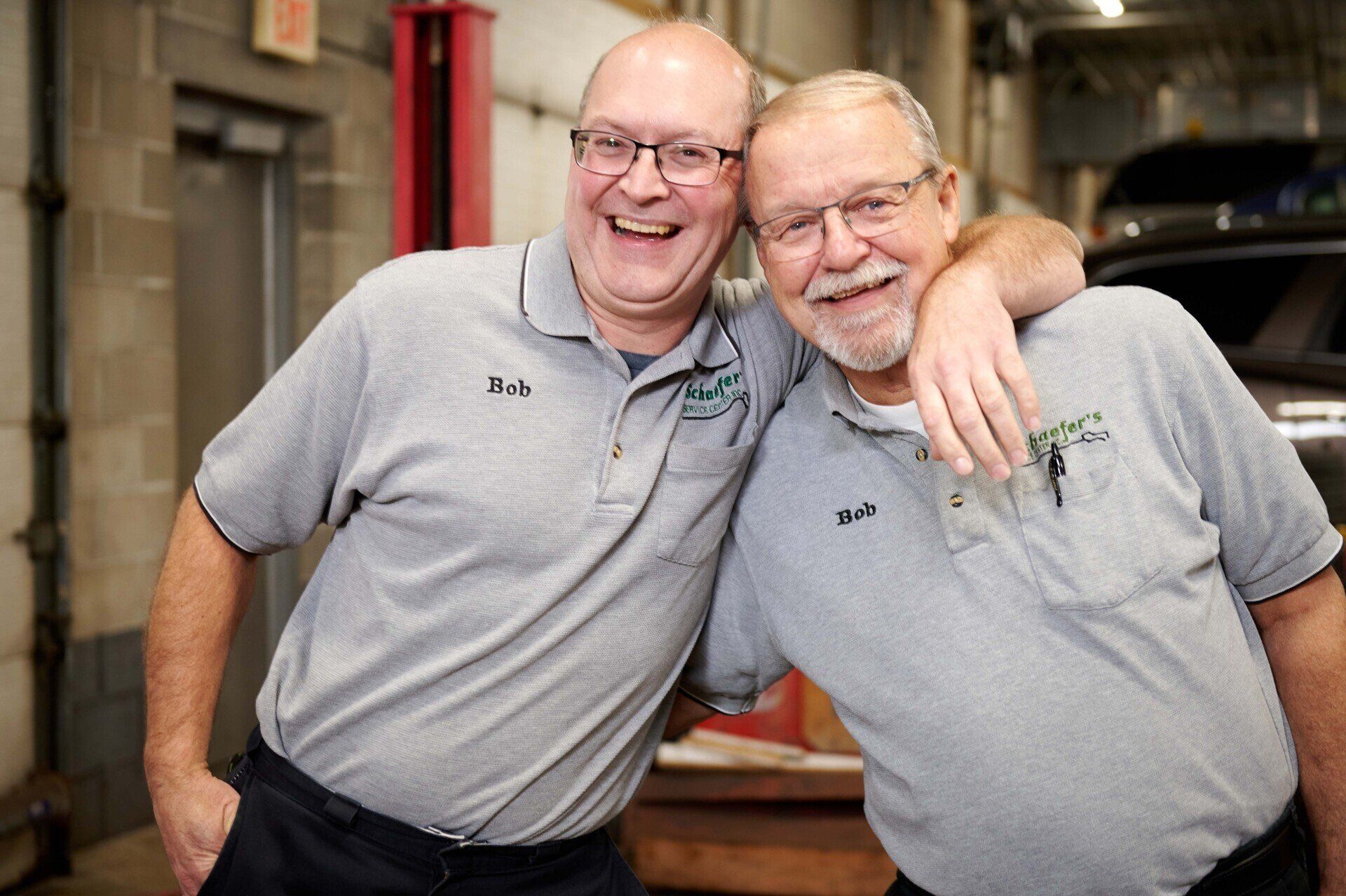 Two men in grey shirts with glasses, smiling and embracing, in a garage.