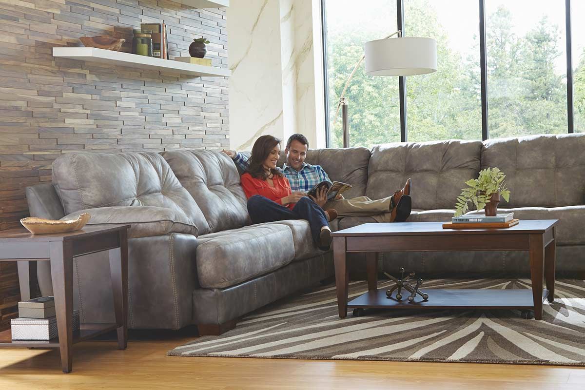 Couple sitting on sectional sofa, looking at tablet, in a living room with large windows and stone wall.