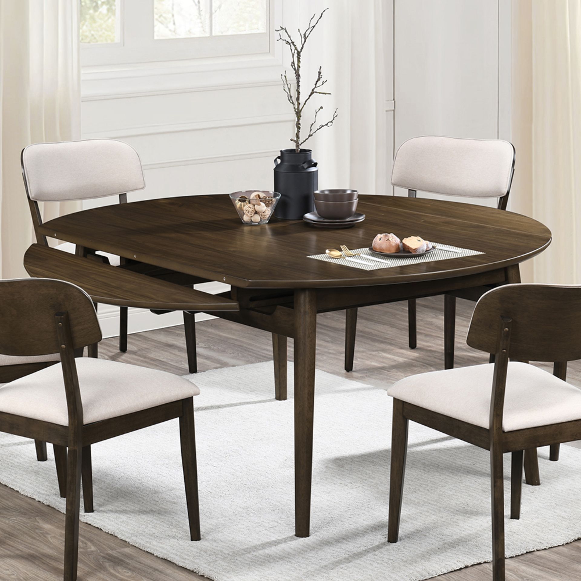 A round, dark wood dining table with two drop leaves partially extended, surrounded by four upholstered chairs on a rug.