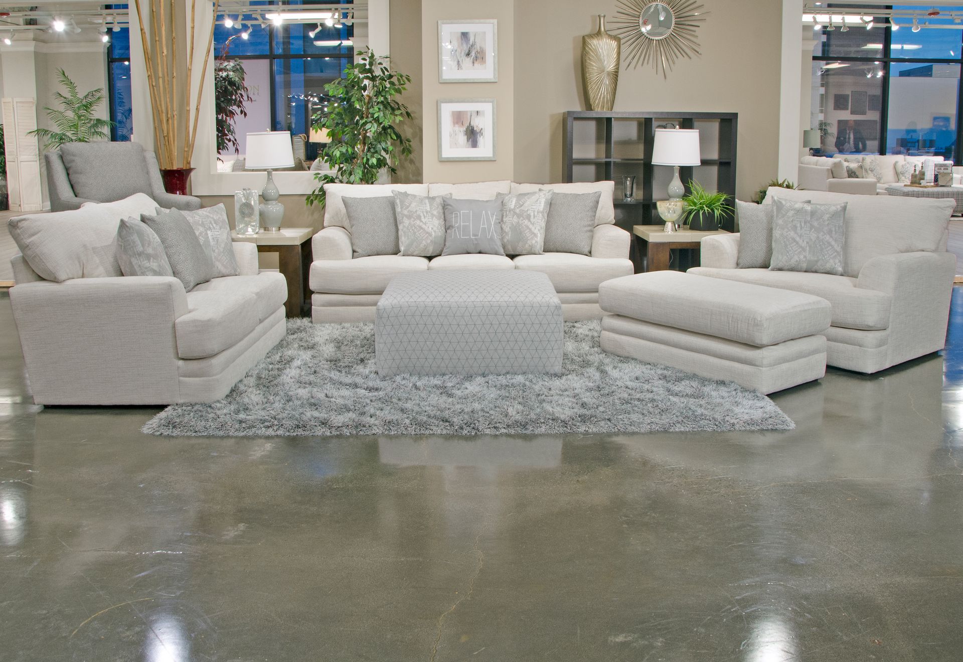 Living room furniture set in a showroom. Beige sofa, chair, ottoman, and rug on a polished concrete floor.