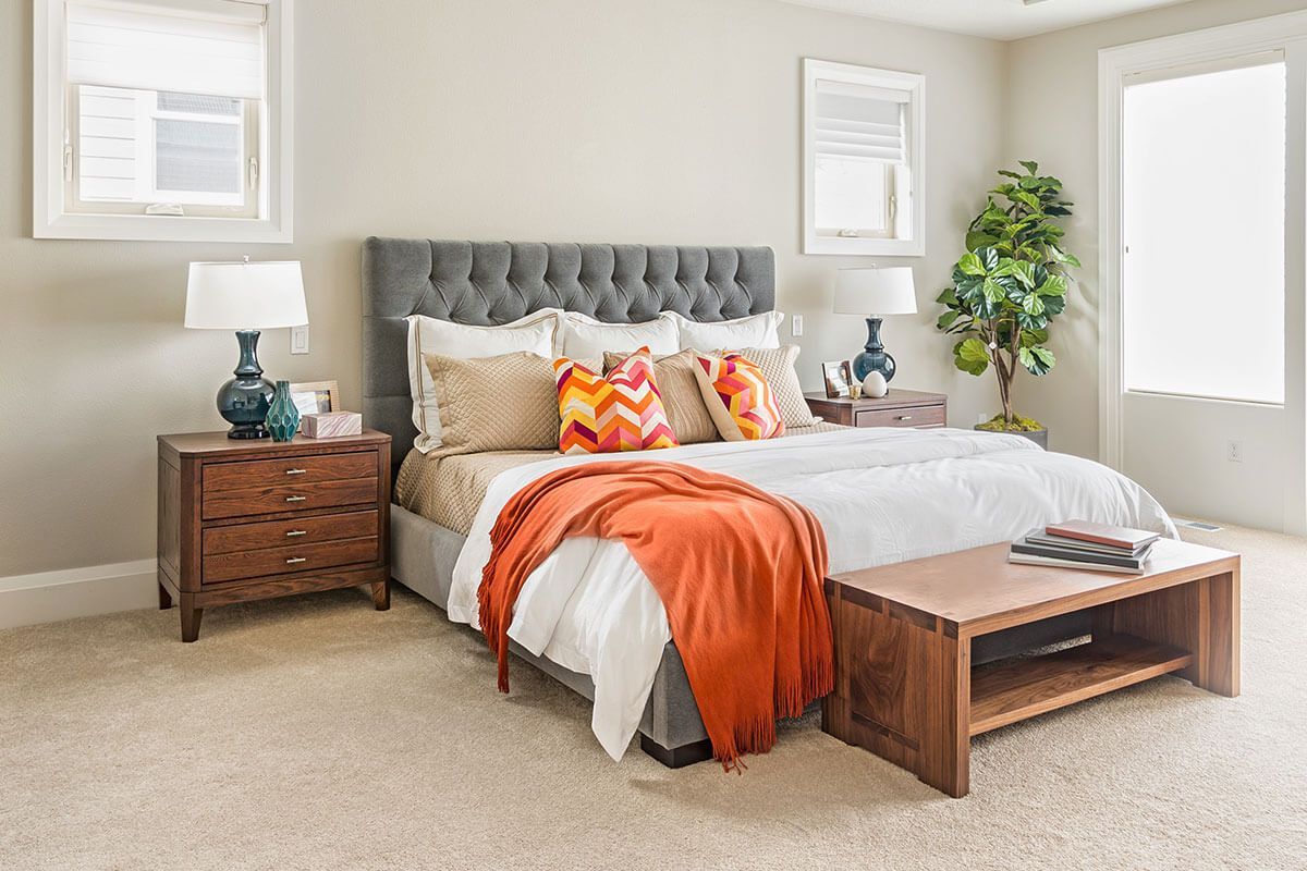 Bedroom with a gray tufted headboard, white bedding, orange accents, and wood furniture.