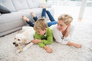 Woman and child lying on a rug, petting a white dog chewing on a bone.