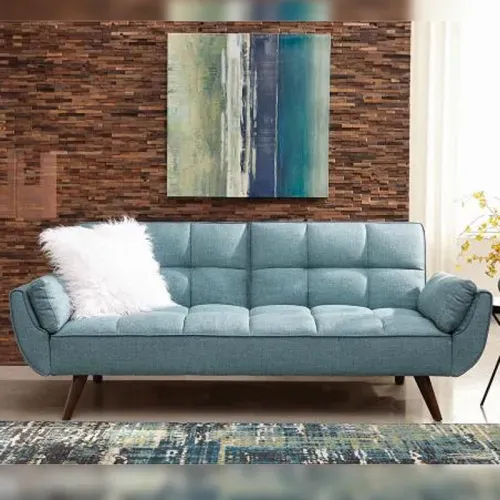 Blue tufted sofa with fluffy white pillow, wooden legs, against a textured brick wall with artwork.