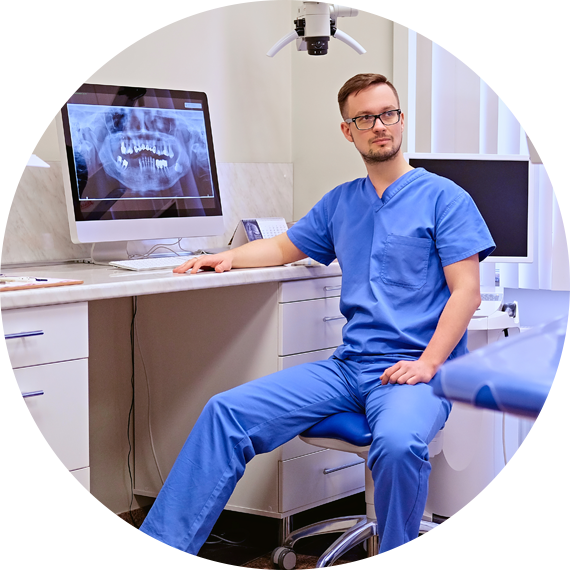 Dentist in blue scrubs seated in a modern dental office beside a computer displaying an X-ray.