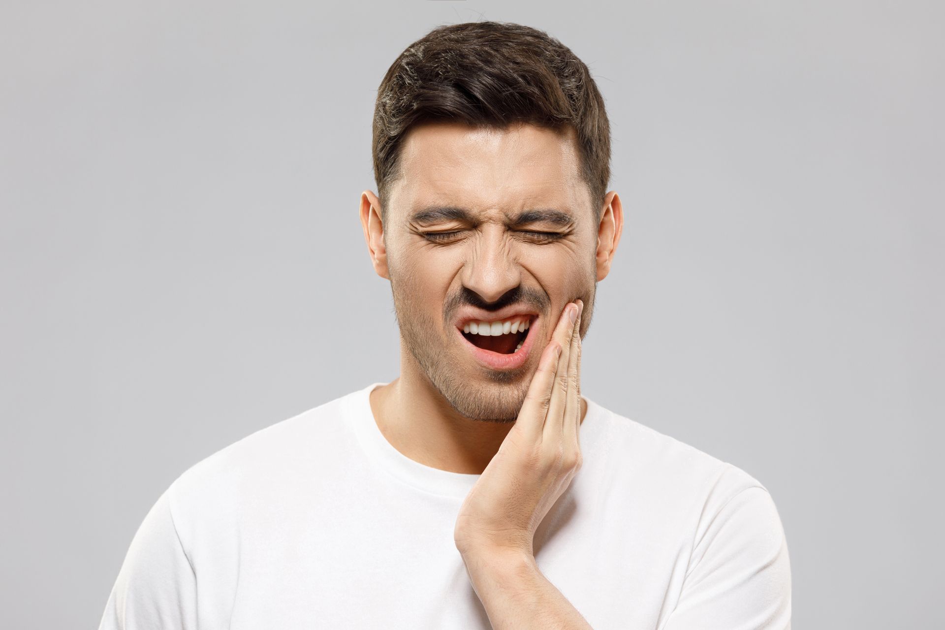 Man in a white shirt grimacing and holding his cheek against a gray background