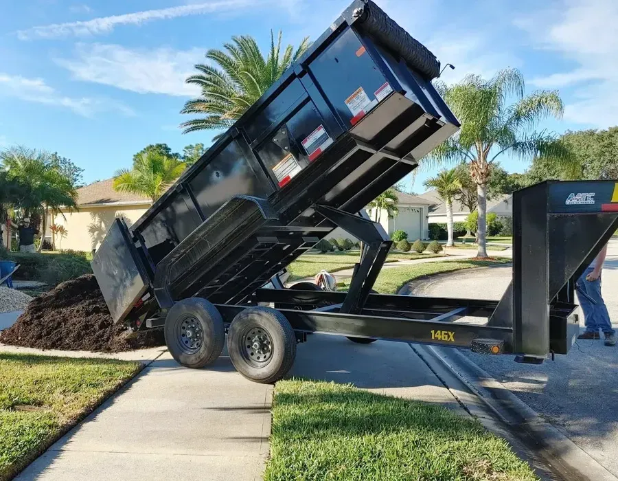 Black dump trailer raised on a driveway, unloading a pile of mulch in a sunny suburban yard.