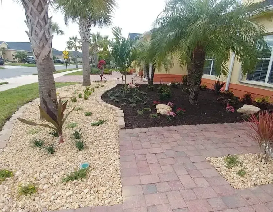 Landscaped front yard with palm trees, white rocks, dark mulch, and a brick walkway by a house