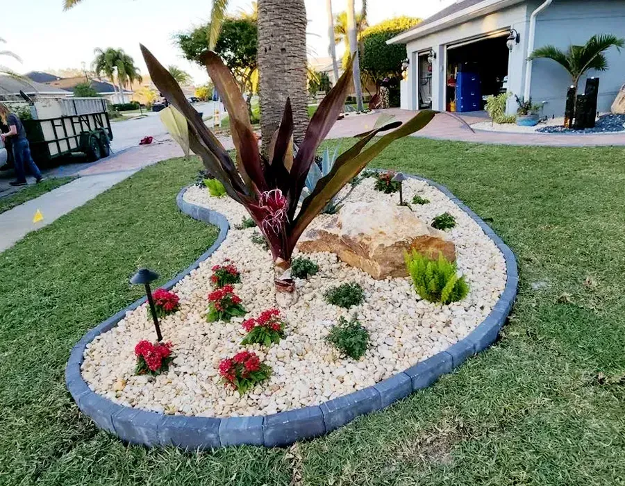 Curved front yard flower bed with white rocks, red flowers, and a tall burgundy plant by a house
