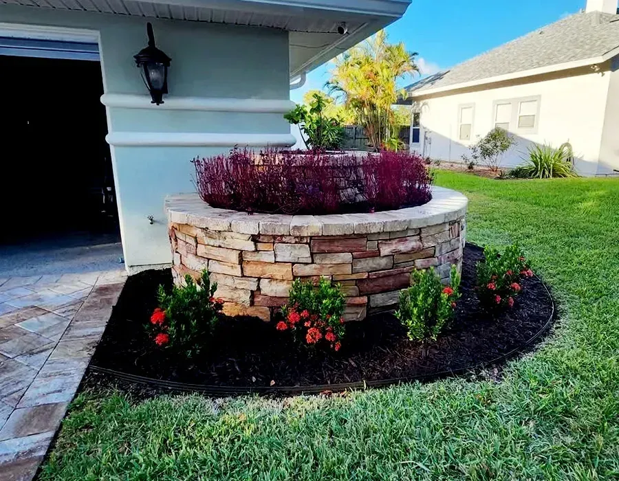 Landscaped stone planter with purple shrubs and red flowers beside a house and lawn