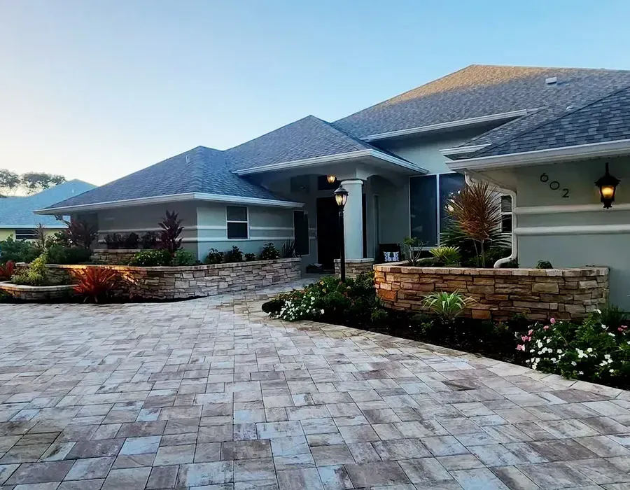 Modern single-story house with a gray shingle roof, stone driveway, and landscaped front yard at dusk