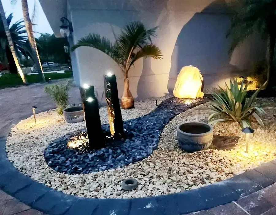Decorative outdoor garden bed with black stones, palm plants, and glowing pathway lights at dusk