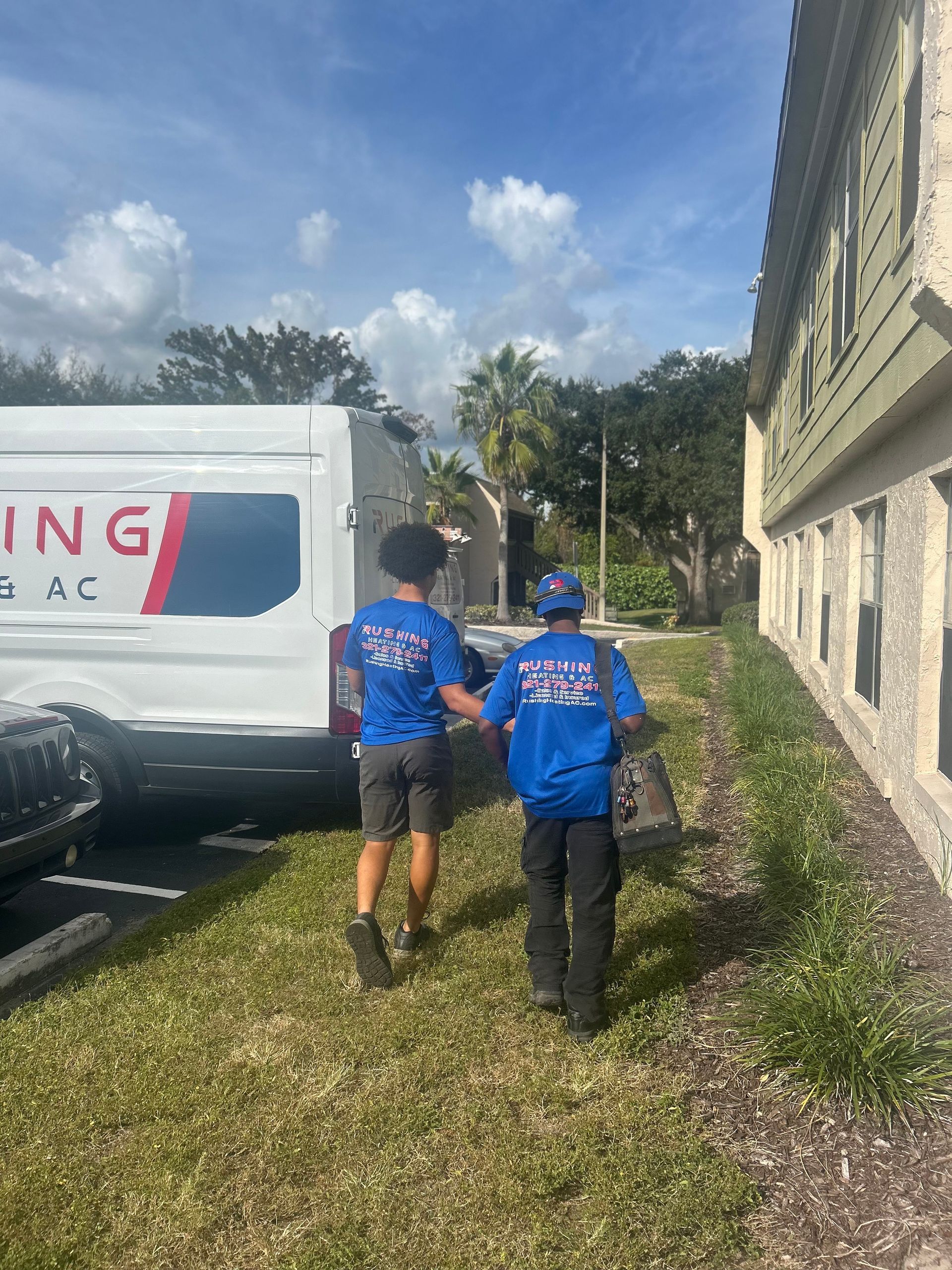 Two people wearing blue shirts are walking alongside a white van. They appear to be carrying equipment near a building.