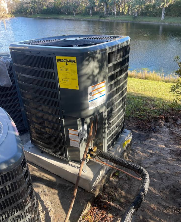 Air conditioning unit outdoors, next to a lake. Gray and black. Yellow energy guide label visible.