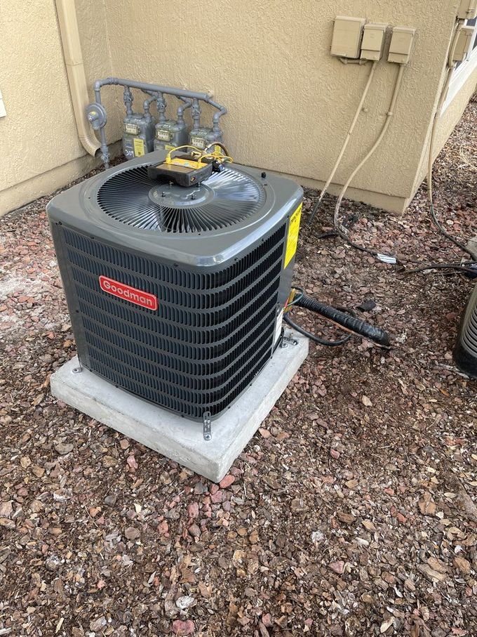 Goodman air conditioning unit on a concrete pad, surrounded by mulch, against a beige building.