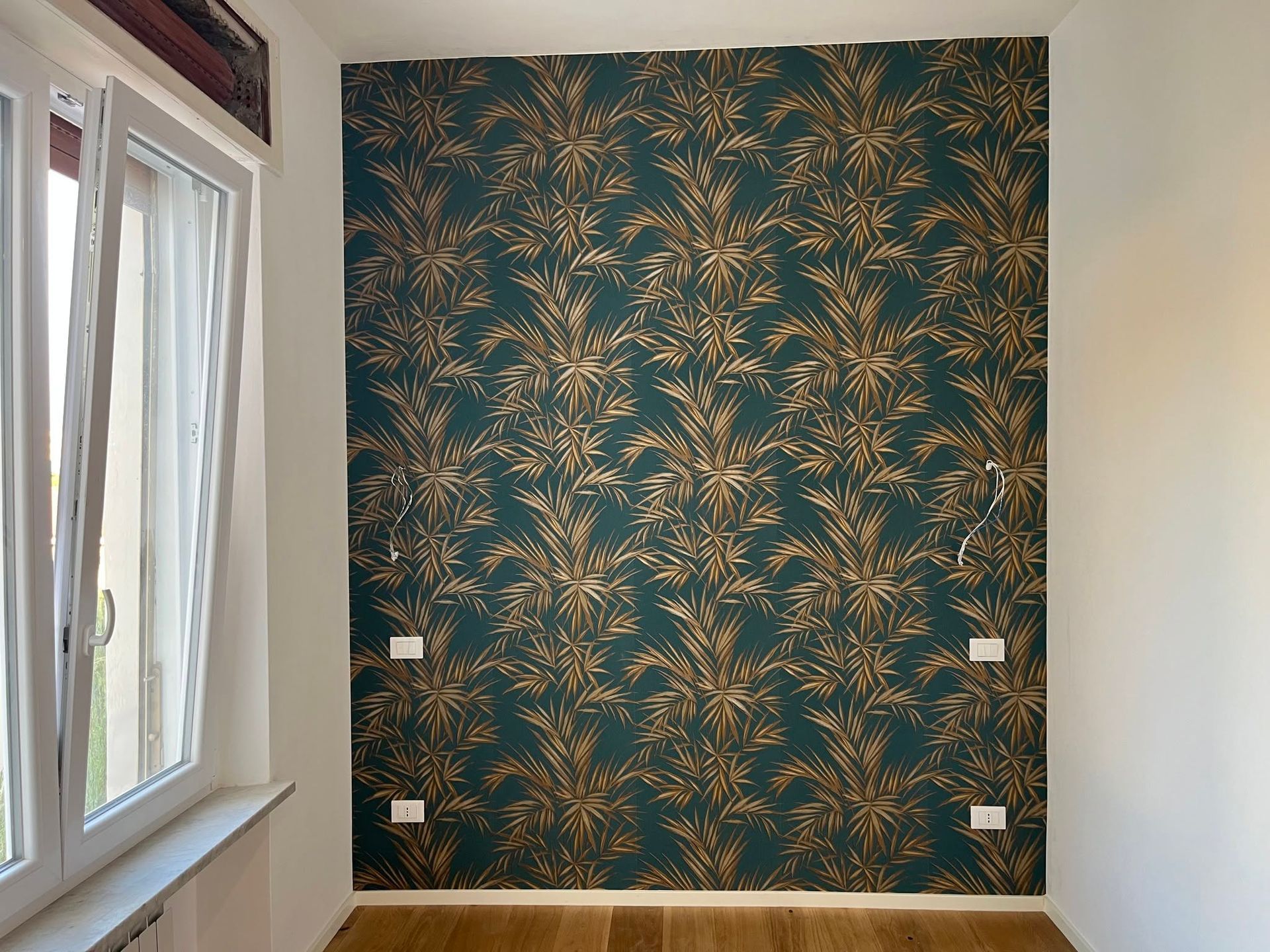 Stanza vuota con una parete decorata con motivi floreali verdi e grandi finestre sulla sinistra.