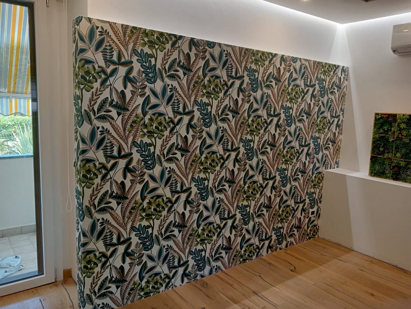 Stanza vuota con carta da parati a foglie tropicali, pavimento in legno chiaro e porta scorrevole in vetro.