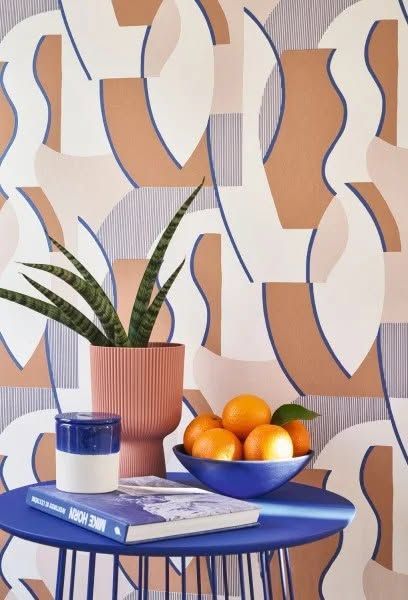 Tavolo blu con una pianta in vaso, una tazza di caffè, un libro e una ciotola di arance su uno sfondo di carta da parati a motivi geometrici.