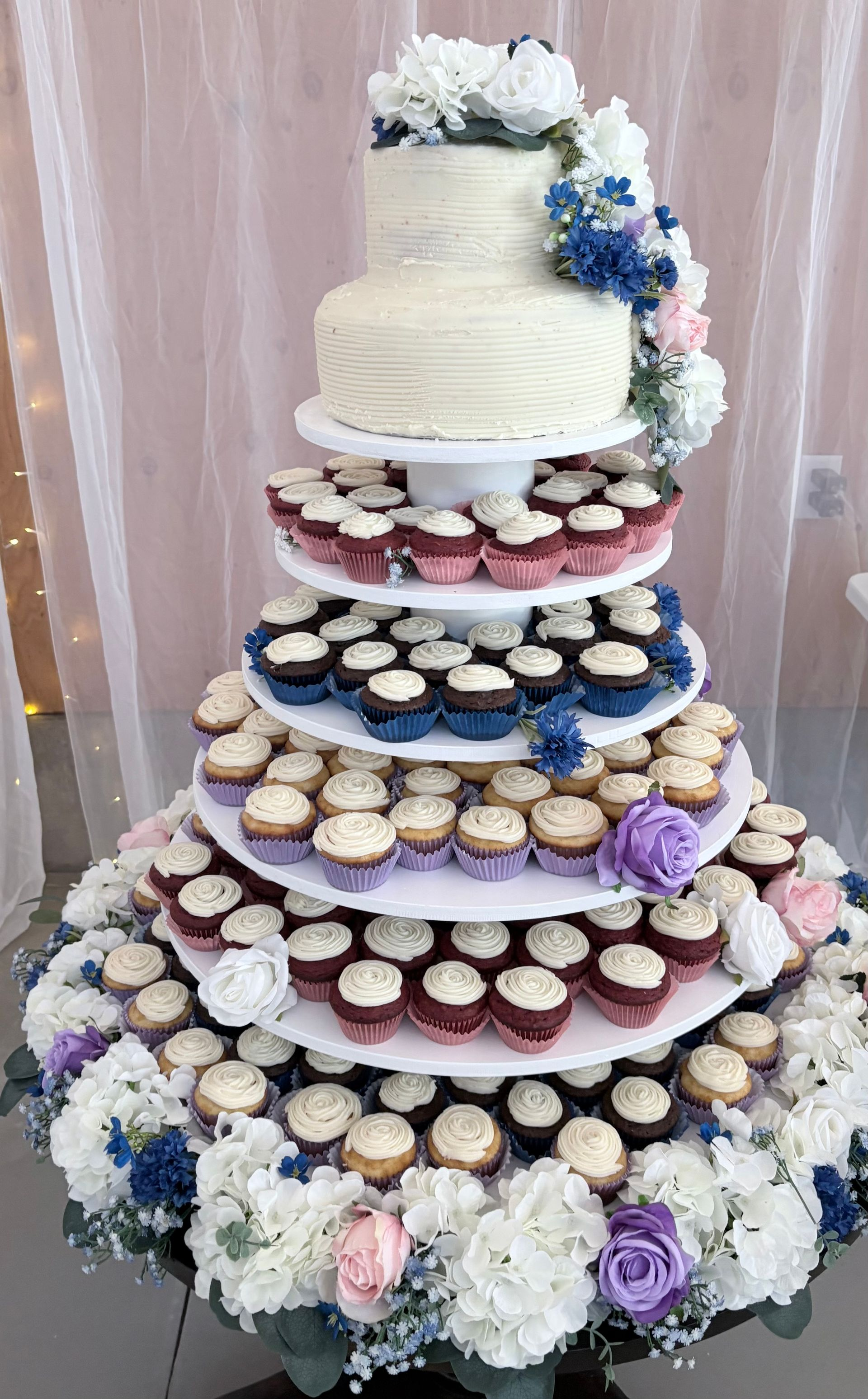 White two-tier wedding cake with roses on a decorated stand, against a backdrop of string lights and flowers