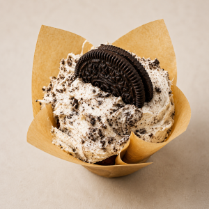 Cookies-and-cream cupcake in a brown paper liner, topped with crushed cookies and an Oreo cookie half