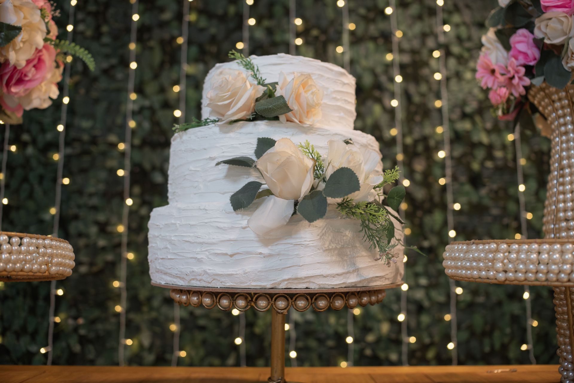 White two-tier wedding cake with roses on a decorated stand, against a backdrop of string lights and flowers