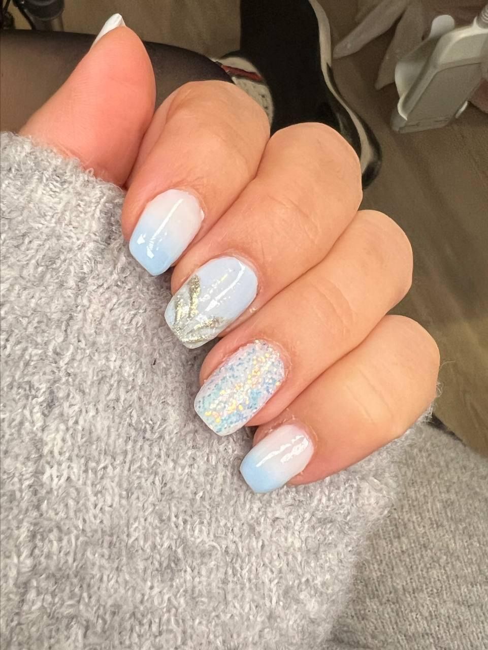 Person's hand with light blue ombre nails; one nail decorated with silver foil and another with glitter.