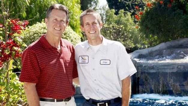 Two men are standing next to each other in front of a waterfall.