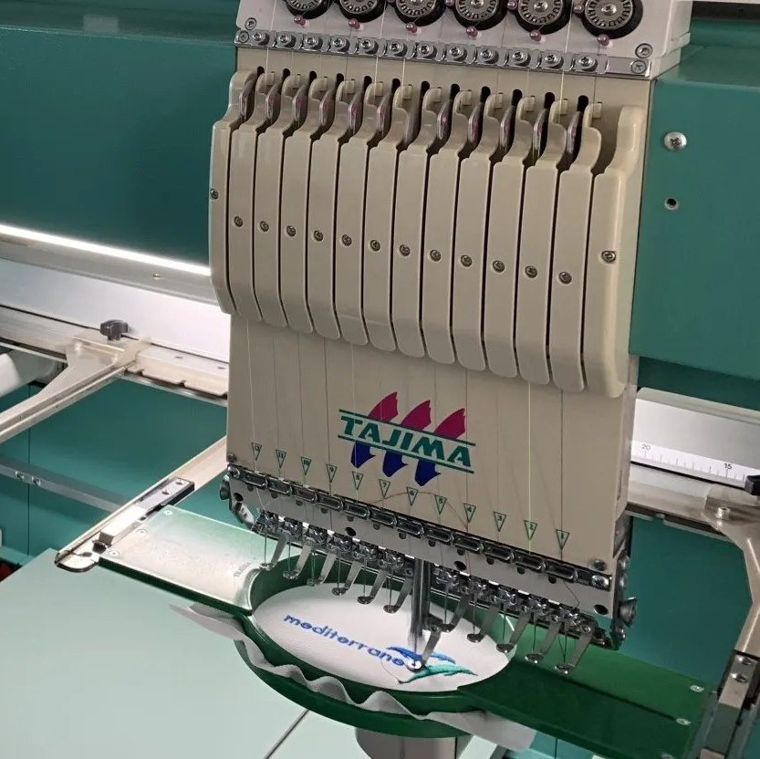 Primo piano di una macchina da ricamo multi-ago Tajima che cuce un capo bianco su un tavolo da lavoro verde.