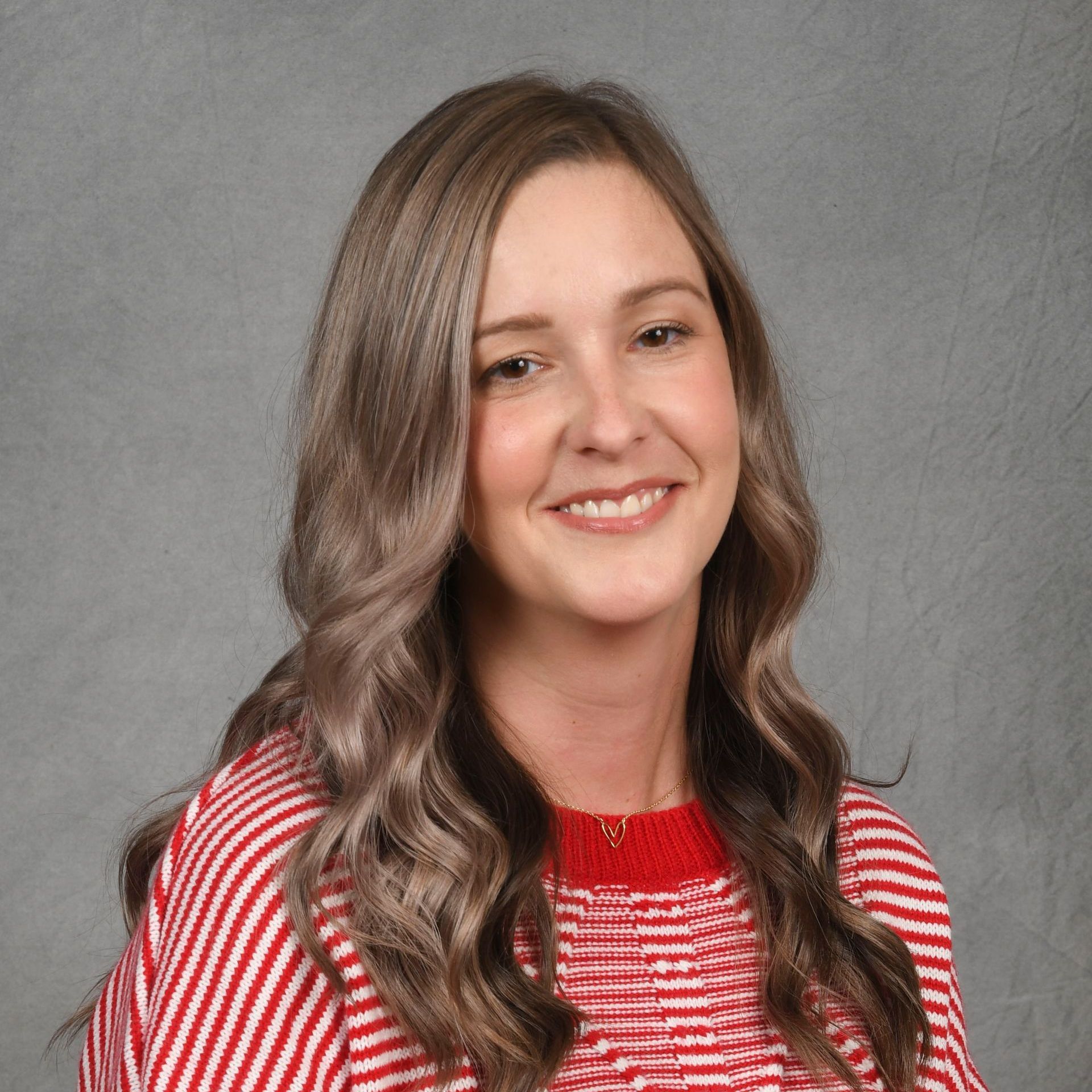 Smiling person with wavy brown hair wearing a red-and-white striped top against a gray background