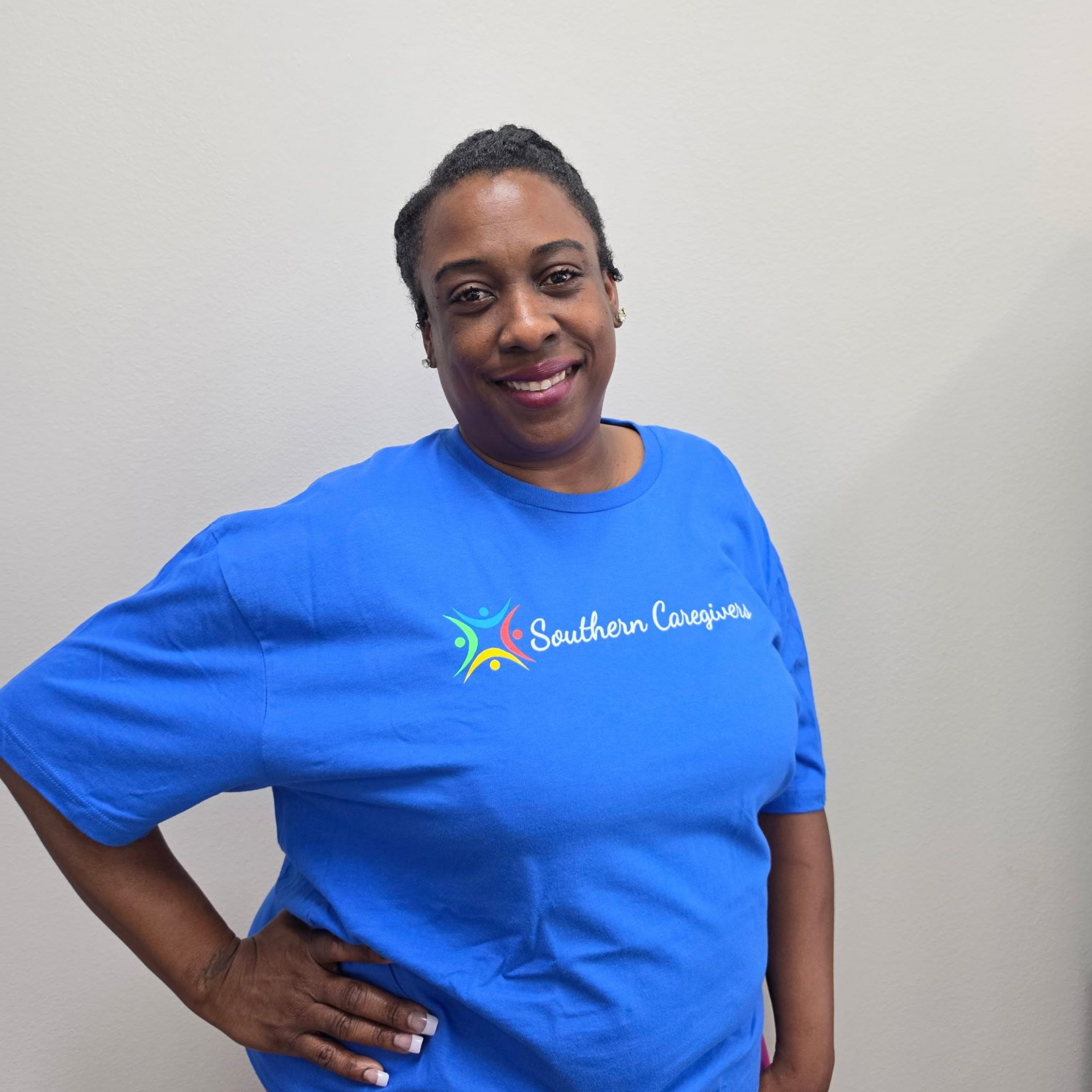 Woman smiling in a blue T-shirt with logo, standing with hand on hip against a plain wall