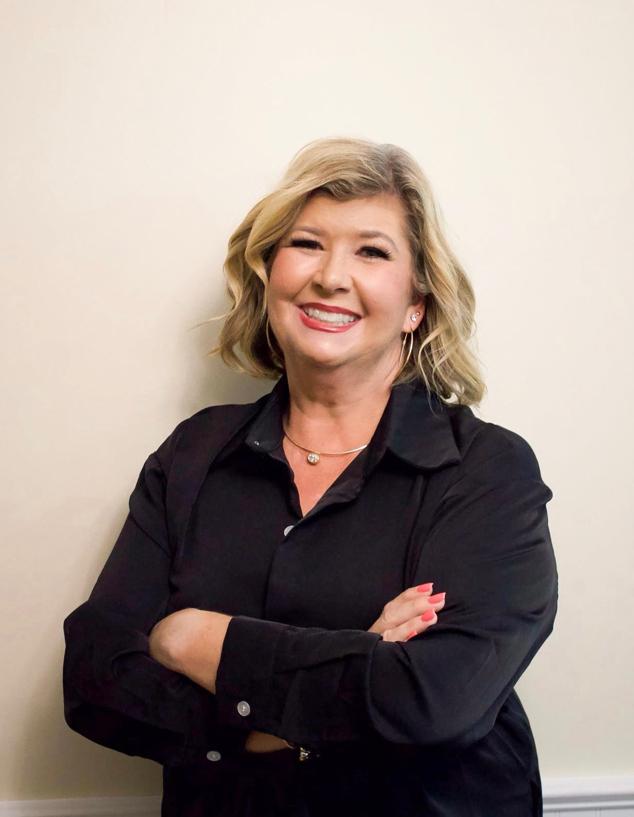 Smiling woman in a black blouse standing with arms crossed against a light wall