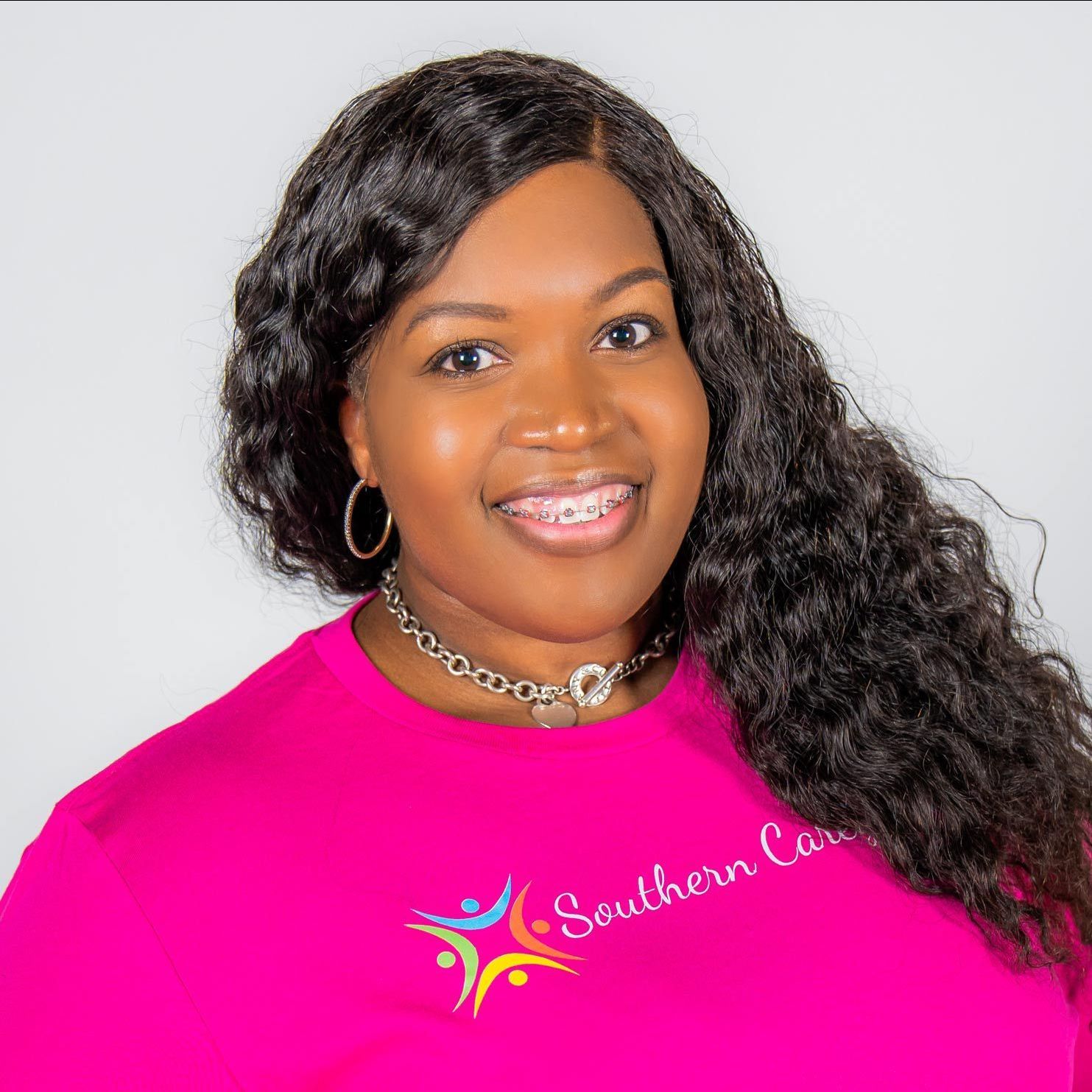 Smiling woman in a bright pink shirt with long curly hair and a necklace against a white background