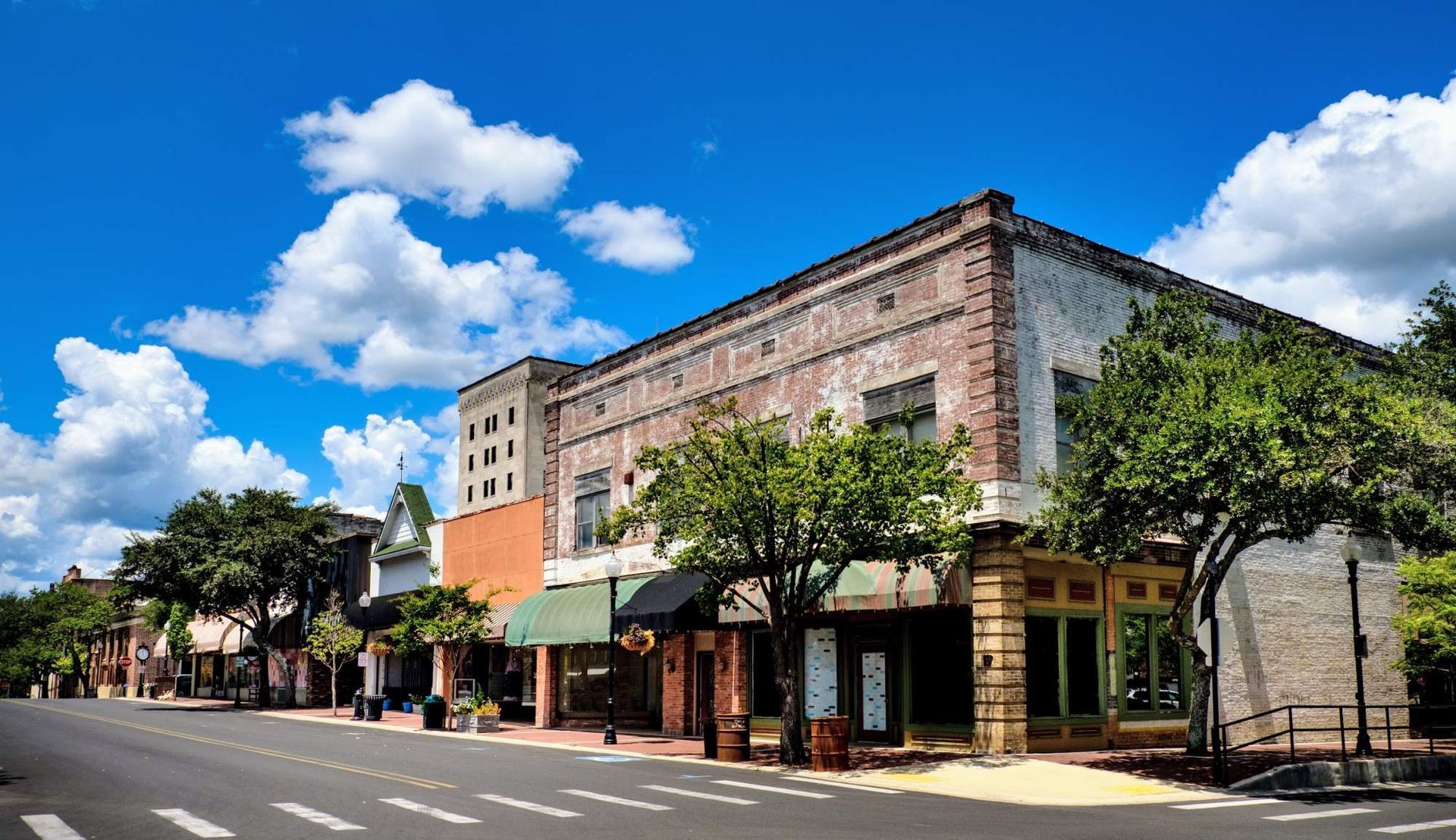 Street with shops under blue sky, trees line the sidewalks, brick and stone buildings.