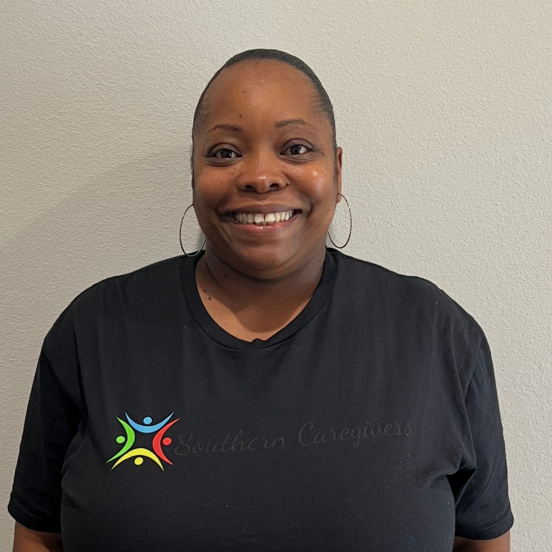 Smiling person in a black T-shirt standing against a plain beige wall.