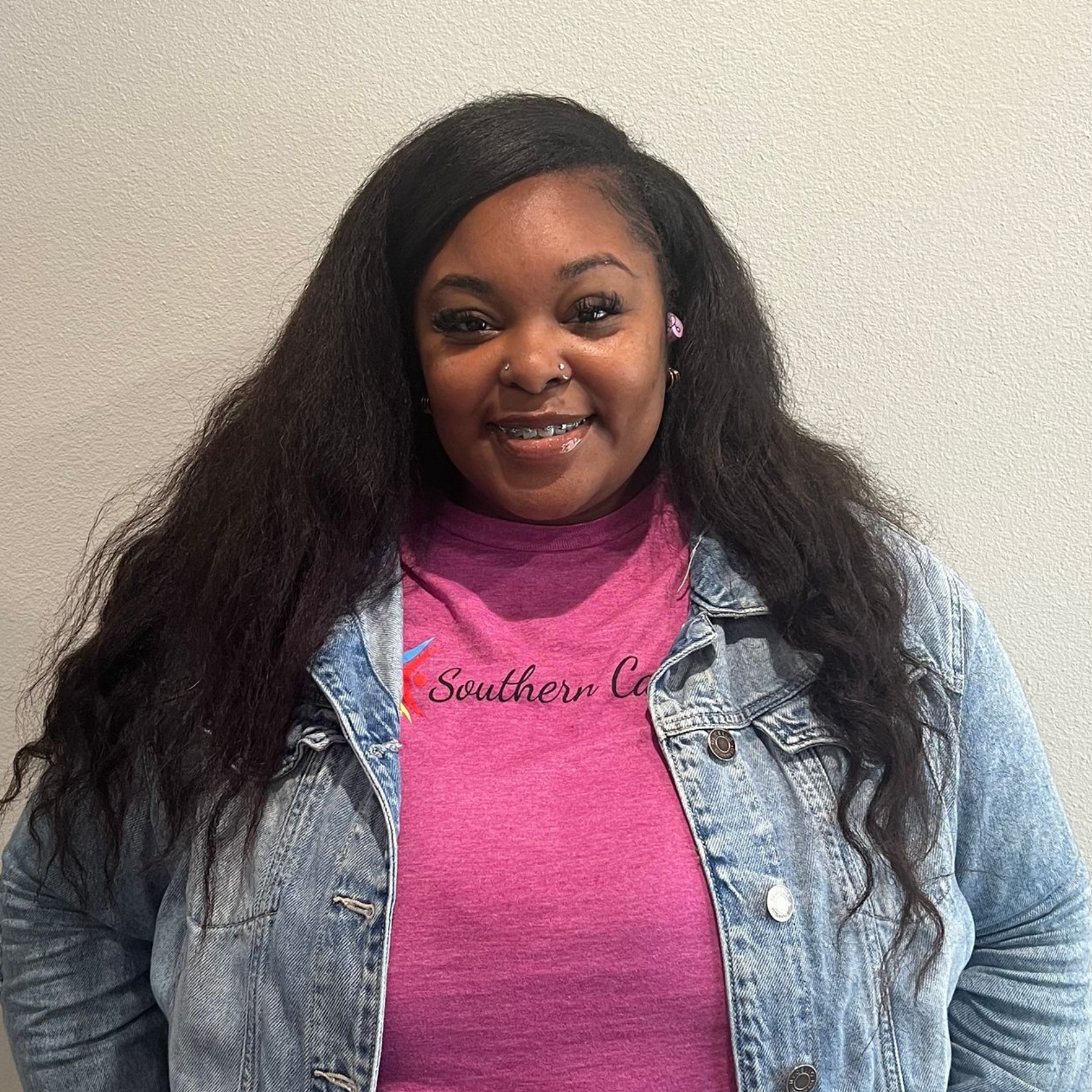 Smiling person in a pink shirt and denim jacket against a plain wall